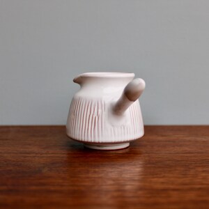 May include: White ceramic creamer with a pink stripe design and a handle shaped like a spout. The creamer is sitting on a wooden surface.