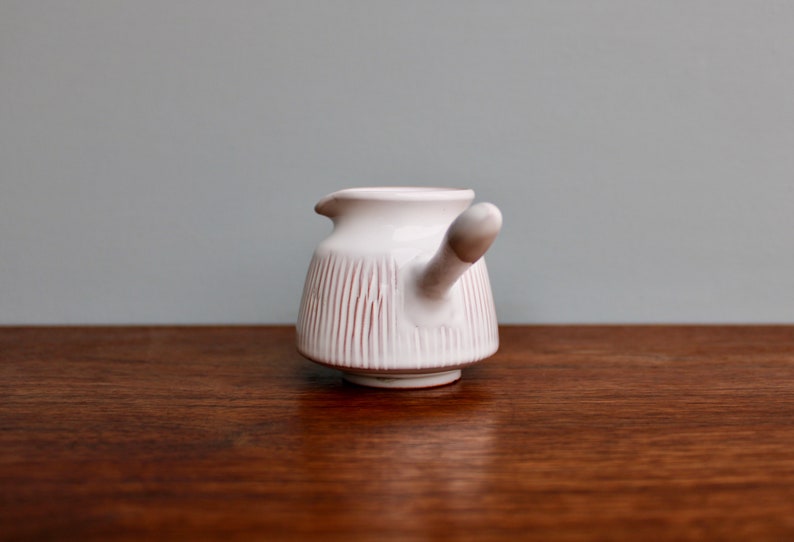May include: White ceramic creamer with a pink stripe design and a handle shaped like a spout. The creamer is sitting on a wooden surface.