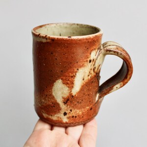May include: A handmade ceramic mug with a brown and white speckled glaze. The mug has a rounded body and a handle.