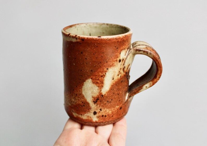 May include: A handmade ceramic mug with a brown and white speckled glaze. The mug has a rounded body and a handle.