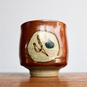 May include: A handmade ceramic cup with a rich brown glaze. The cup features a beige circular design with a dark blue and brown abstract pattern. The cup has a rounded body and a slightly flared base, perfect for enjoying a warm beverage.