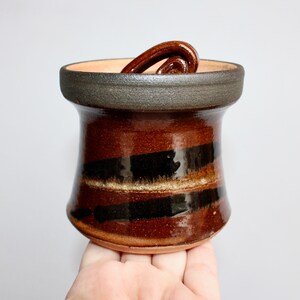 May include: A handmade ceramic jar with a dark brown and black striped design. The jar has a dark rim and a decorative handle. The jar is approximately 4 inches tall and 3 inches wide.