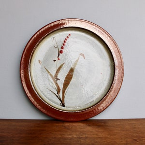 May include: A round ceramic plate with a brown rim and a white center. The plate features a hand-painted design of a stylized flower with red berries and brown leaves.