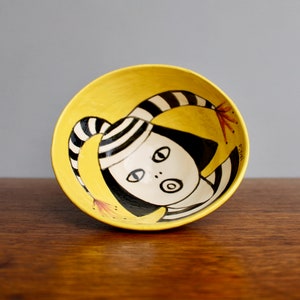 May include: A yellow ceramic bowl with a hand-painted design of a person with black and white striped hat and clothing. The person has large eyes and an open mouth. The bowl is on a wooden surface.