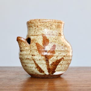 May include: A small, brown and white ceramic pitcher with a brown leaf design. The pitcher has a spout and a handle.