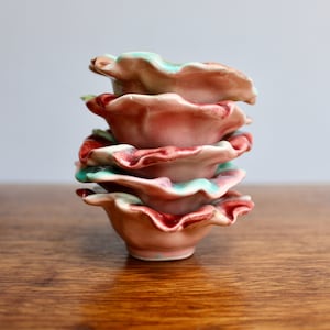 May include: A stack of four small, ceramic bowls with a ruffled edge. The bowls are a pale pink with a red and turquoise trim. The bowls are stacked on top of each other on a wooden surface.
