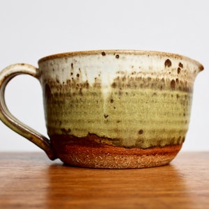 May include: A brown and white ceramic pitcher with a green glaze and a brown handle. The pitcher has a rustic, handmade look.