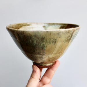 May include: A handmade ceramic bowl with a wide, angled rim. The bowl features a blend of earthy tones, including greens, browns, and creams. The bowl is held up by a hand, showcasing its shape and size.