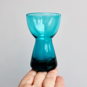 May include: A small, teal blue glass vase with a wide mouth and a narrow base. The vase is held in a hand.