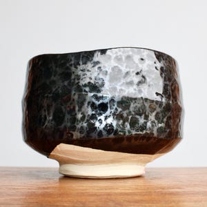 May include: A handcrafted ceramic bowl with a textured, dark, speckled exterior. The bowl has a wide, irregular rim and a light-colored base. The interior appears to be glazed with a glossy finish. The bowl is sitting on a wooden surface.