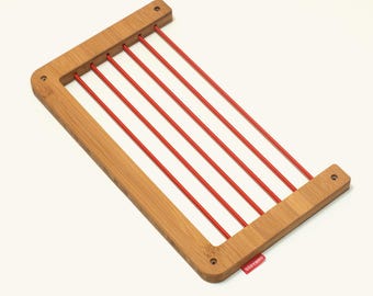 May include: A rectangular bamboo frame featuring five parallel red elastic cords stretched across the center. The frame has four small circular holes at the corners for mounting and a small red fabric tag with the word ONWARDS in white.