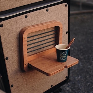 May include: A bamboo fold-out shelf with a built-in cup holder and a small paper cup. The shelf is mounted on a textured, light brown surface. The cup has a logo and a wooden stirrer.