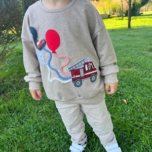 Könnte beinhalten: Ein beiges Sweatshirt mit einem farbenfrohen Applikation eines roten Feuerwehrautos, eines roten Ballons und eines blauen Autos. Das Sweatshirt wird mit beiger Hose und weißen Turnschuhen mit schwarzen Details getragen. Das Kind steht auf grünem Gras.