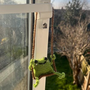 May include: A handmade, green crochet frog on a swing. The frog has black eyes and a smiling mouth. The swing is made of brown yarn and is hanging from a window handle. The background is a blurred view of a sunny day.
