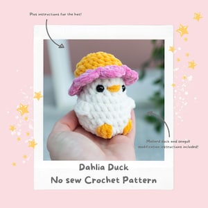 Op de afbeelding: Een handgemaakte witte gehaakte eend met een gele en roze hoed. De eend heeft zwarte ogen, een oranje snavel en voeten. De tekst "Dahlia Duck No sew Crochet Pattern" staat onderaan de afbeelding. Instructies voor de hoed en aanpassingen zijn inbegrepen.