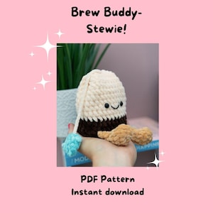 Puede incluir: Un peluche de ganchillo con forma de taza de café con una cara sonriente. El juguete es principalmente de color crema con una base marrón y un asa beige. También se ve el texto "Brew Buddy-Stewie!" y "PDF Pattern Instant download".