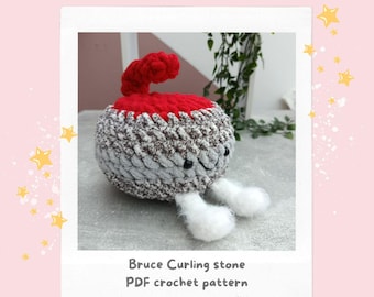 Pode incluir: Uma pedra de curling de crochê com uma alça vermelha e um corpo cinza e branco. A pedra tem um rosto sorridente e pés brancos. A imagem inclui o texto "Crochet Pattern" e "Bruce Curling stone PDF crochet pattern."