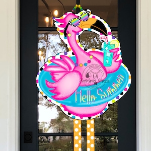 May include: Pink flamingo wearing sunglasses and a float holding a glass of lemonade with a lemon wedge. The flamingo has yellow and white polka dot legs and is wearing purple and green flip flops. The float says "Hello Summer".