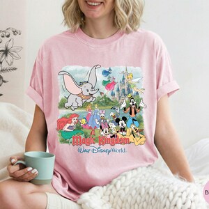 May include: A light pink short-sleeve t-shirt featuring a graphic print of various Disney characters including Dumbo, Mickey Mouse, and Tinker Bell in front of a castle, with the text Magic Kingdom Walt Disney World printed below the characters.