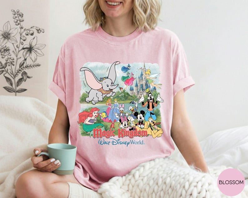 May include: A light pink short-sleeve t-shirt featuring a graphic print of various Disney characters including Dumbo, Mickey Mouse, and Tinker Bell in front of a castle, with the text Magic Kingdom Walt Disney World printed below the characters.