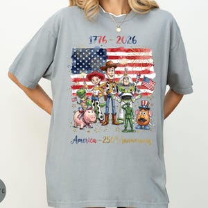 Pu&ograve; includere: T-shirt grigio chiaro con una grafica della bandiera americana, fuochi d'artificio e personaggi dei cartoni animati. Il testo "1776-2026" e "America - 250th anniversary" sono stampati. La maglietta ha una vestibilit&agrave; ampia.