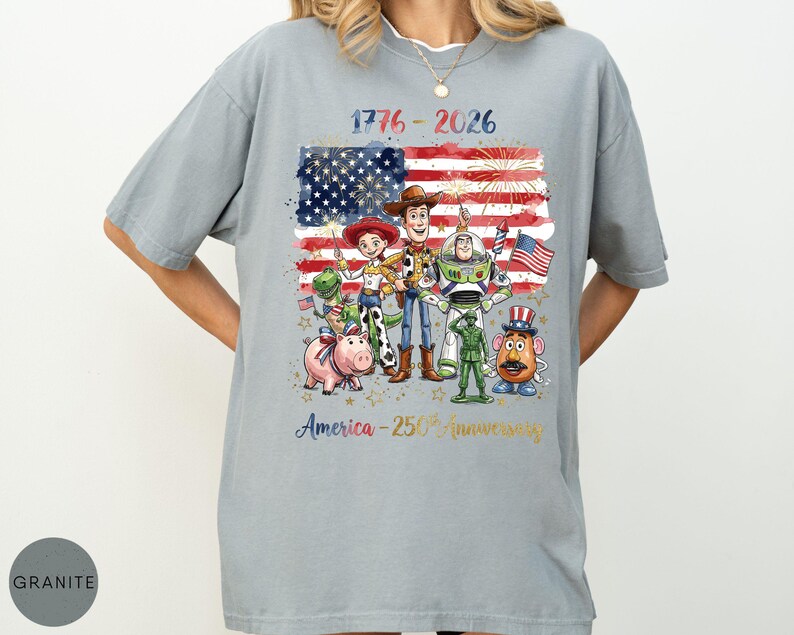 Pu&ograve; includere: T-shirt grigio chiaro con una grafica della bandiera americana, fuochi d'artificio e personaggi dei cartoni animati. Il testo "1776-2026" e "America - 250th anniversary" sono stampati. La maglietta ha una vestibilit&agrave; ampia.