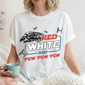 May include: A white short-sleeved t-shirt featuring a graphic of a spaceship and TIE fighters. The text reads RED WHITE AND PEW PEW PEW in red, white, and blue colors. The design includes a blue rectangular outline surrounding the word WHITE.