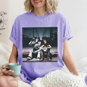 May include: A light purple short-sleeve t-shirt featuring a graphic print of the characters Buzz Lightyear and Woody sitting together on a curb at night. The design shows the two figures in a dark, urban setting with utility poles in the background.