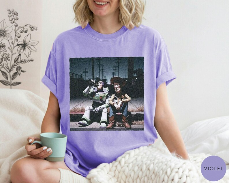 May include: A light purple short-sleeve t-shirt featuring a graphic print of the characters Buzz Lightyear and Woody sitting together on a curb at night. The design shows the two figures in a dark, urban setting with utility poles in the background.