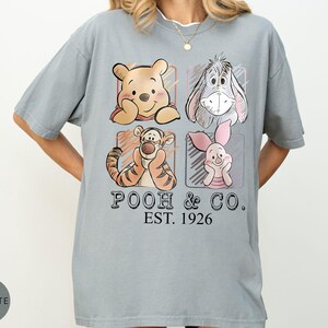 May include: A light gray t-shirt featuring four cartoon characters: Winnie the Pooh, Eeyore, Tigger, and Piglet. The text "POOH & CO. EST. 1926" is printed below the characters. The shirt has short sleeves and a relaxed fit.