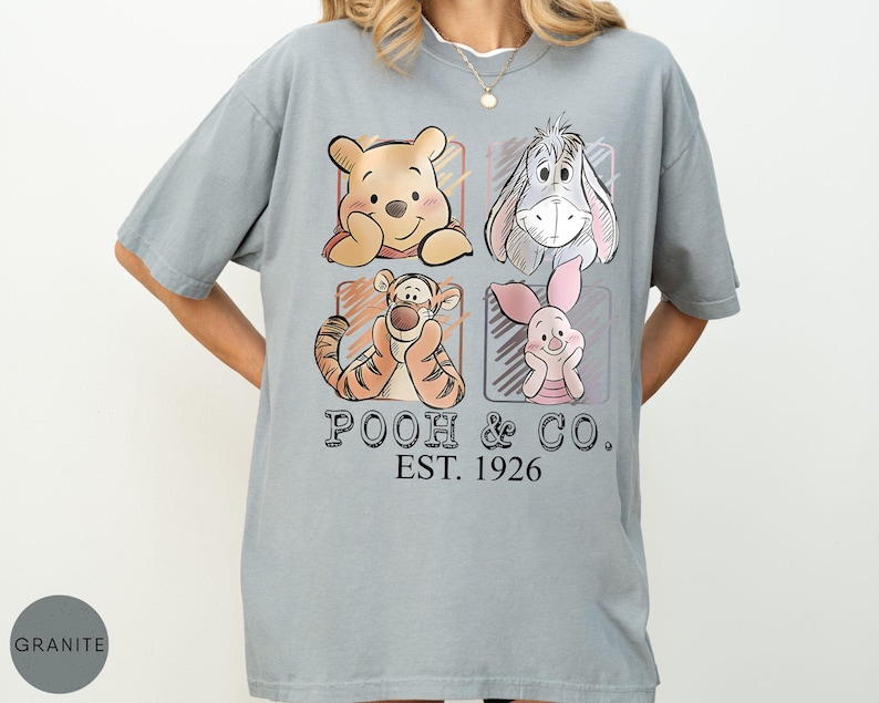 May include: A light gray t-shirt featuring four cartoon characters: Winnie the Pooh, Eeyore, Tigger, and Piglet. The text "POOH & CO. EST. 1926" is printed below the characters. The shirt has short sleeves and a relaxed fit.