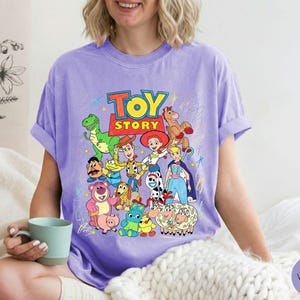 May include: A light purple t-shirt featuring a colourful graphic print of various Toy Story characters including Woody, Buzz Lightyear, Jessie, Rex, and others, with the Toy Story logo at the top and scattered crayon-like scribbles in the background.