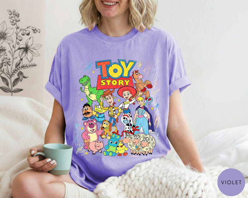 May include: A light purple t-shirt featuring a colourful graphic print of various Toy Story characters including Woody, Buzz Lightyear, Jessie, Rex, and others, with the Toy Story logo at the top and scattered crayon-like scribbles in the background.