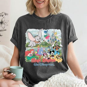 May include: A charcoal gray short-sleeve t-shirt featuring a graphic print of Dumbo, Tinker Bell, Ariel, Mickey Mouse, and other characters in front of a castle. The text Magic Kingdom and Walt Disney World is printed in red and blue below the characters.
