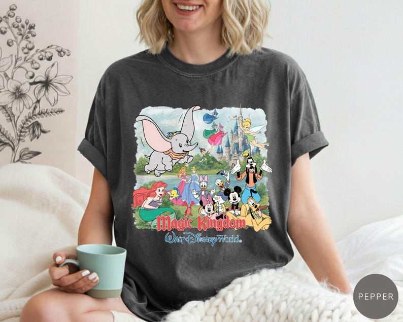 May include: A charcoal gray short-sleeve t-shirt featuring a graphic print of Dumbo, Tinker Bell, Ariel, Mickey Mouse, and other characters in front of a castle. The text Magic Kingdom and Walt Disney World is printed in red and blue below the characters.