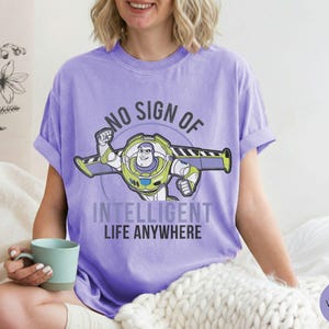 Puede incluir: Una camiseta violeta con un gr&aacute;fico de Buzz Lightyear y el texto "NO SIGN OF INTELLIGENT LIFE ANYWHERE". El dise&ntilde;o presenta a Buzz en su traje espacial con detalles verdes, amarillos y blancos. La camiseta es de estilo casual.