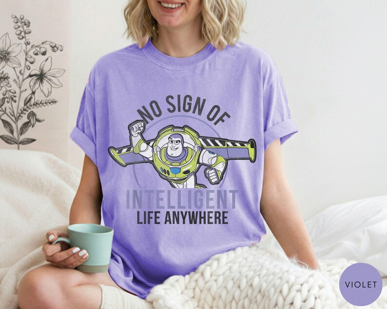 Puede incluir: Una camiseta violeta con un gr&aacute;fico de Buzz Lightyear y el texto "NO SIGN OF INTELLIGENT LIFE ANYWHERE". El dise&ntilde;o presenta a Buzz en su traje espacial con detalles verdes, amarillos y blancos. La camiseta es de estilo casual.