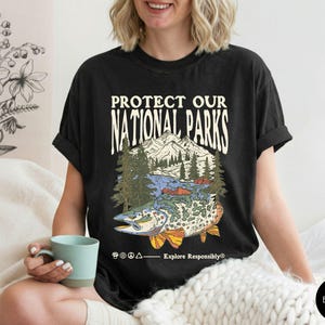 May include: Black t-shirt with a graphic that reads "PROTECT OUR NATIONAL PARKS." The graphic features a mountain range, trees, a lake, and a fish. The shirt also has the text "Explore Responsibly&reg;".