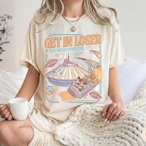 Pode incluir: Camiseta cor marfim com um design gr&aacute;fico retr&ocirc; com o texto "GET IN LOSER WE'RE GOING TO SPACE". O gr&aacute;fico inclui um passeio de parque tem&aacute;tico espacial com personagens de desenhos animados, planetas e OVNIs. A camiseta tem um ajuste folgado.