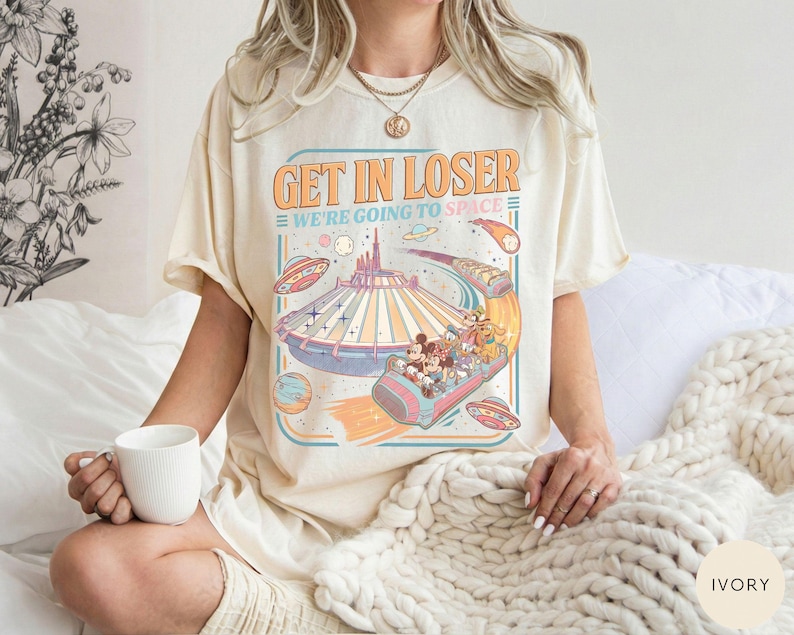 Pode incluir: Camiseta cor marfim com um design gr&aacute;fico retr&ocirc; com o texto "GET IN LOSER WE'RE GOING TO SPACE". O gr&aacute;fico inclui um passeio de parque tem&aacute;tico espacial com personagens de desenhos animados, planetas e OVNIs. A camiseta tem um ajuste folgado.