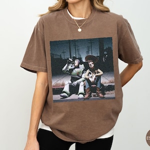 May include: A brown short-sleeve t-shirt featuring a graphic print of the characters Buzz Lightyear and Woody sitting on a curb at night. The shirt has a crew neckline and a relaxed fit, showcasing a detailed scene with utility poles.