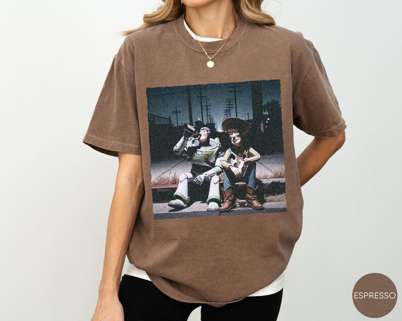 May include: A brown short-sleeve t-shirt featuring a graphic print of the characters Buzz Lightyear and Woody sitting on a curb at night. The shirt has a crew neckline and a relaxed fit, showcasing a detailed scene with utility poles.