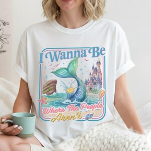 May include: White t-shirt with a colorful graphic featuring a mermaid tail, castle, and the text "I Wanna Be Where The People Aren't." The design includes a blue, pink, and yellow color scheme, evoking a whimsical, beachy vibe. The shirt is a casual, everyday wear item.