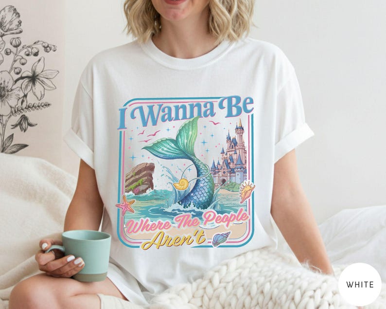 May include: White t-shirt with a colorful graphic featuring a mermaid tail, castle, and the text "I Wanna Be Where The People Aren't." The design includes a blue, pink, and yellow color scheme, evoking a whimsical, beachy vibe. The shirt is a casual, everyday wear item.