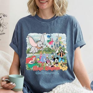 May include: A blue denim-colored t-shirt featuring a graphic print of various Disney characters including Dumbo, Ariel, Mickey Mouse, and Goofy in front of a castle. The text "Magic Kingdom" and "Walt Disney World" is printed below the characters.