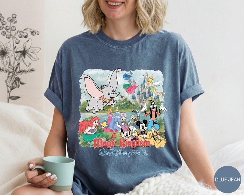 May include: A blue denim-colored t-shirt featuring a graphic print of various Disney characters including Dumbo, Ariel, Mickey Mouse, and Goofy in front of a castle. The text "Magic Kingdom" and "Walt Disney World" is printed below the characters.
