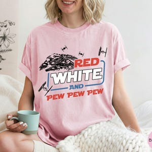May include: A light pink short-sleeve t-shirt featuring a black graphic of a spaceship and smaller fighter crafts. The text reads RED WHITE AND PEW PEW PEW in red and white block lettering, framed by a blue rectangular outline on the chest.