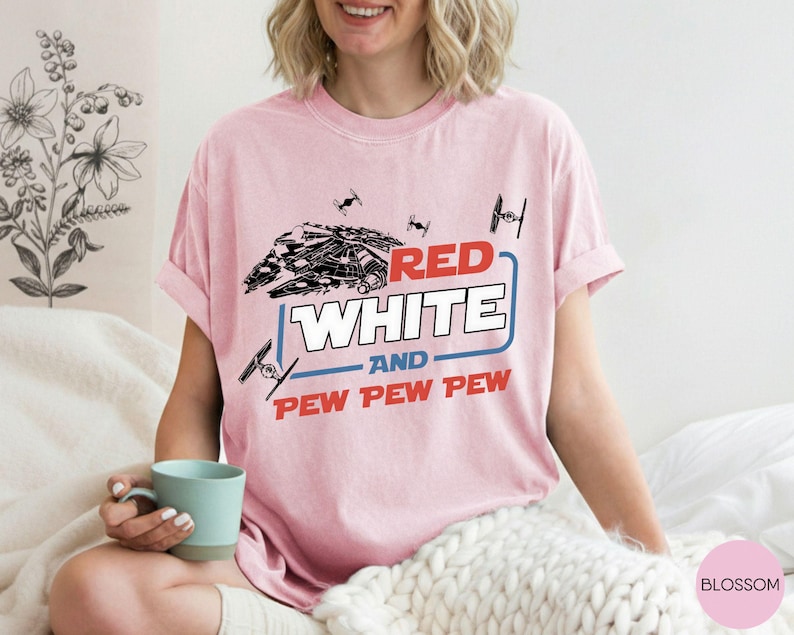 May include: A light pink short-sleeve t-shirt featuring a black graphic of a spaceship and smaller fighter crafts. The text reads RED WHITE AND PEW PEW PEW in red and white block lettering, framed by a blue rectangular outline on the chest.