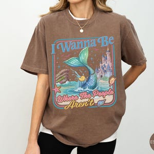 May include: Espresso-colored t-shirt featuring a mermaid illustration with the text "I Wanna Be Where The People Aren't." The design includes a mermaid, castle, and ocean elements in a retro style. The shirt is a relaxed fit.