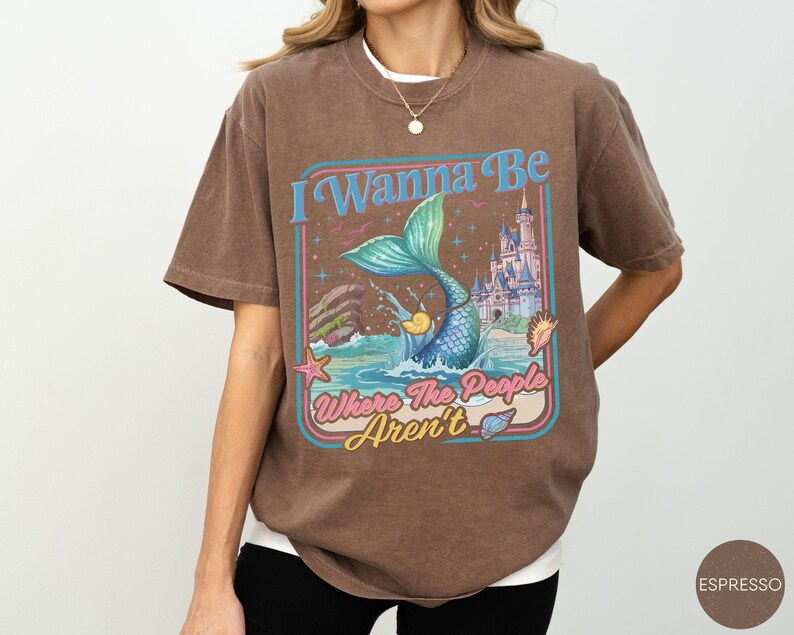 May include: Espresso-colored t-shirt featuring a mermaid illustration with the text "I Wanna Be Where The People Aren't." The design includes a mermaid, castle, and ocean elements in a retro style. The shirt is a relaxed fit.